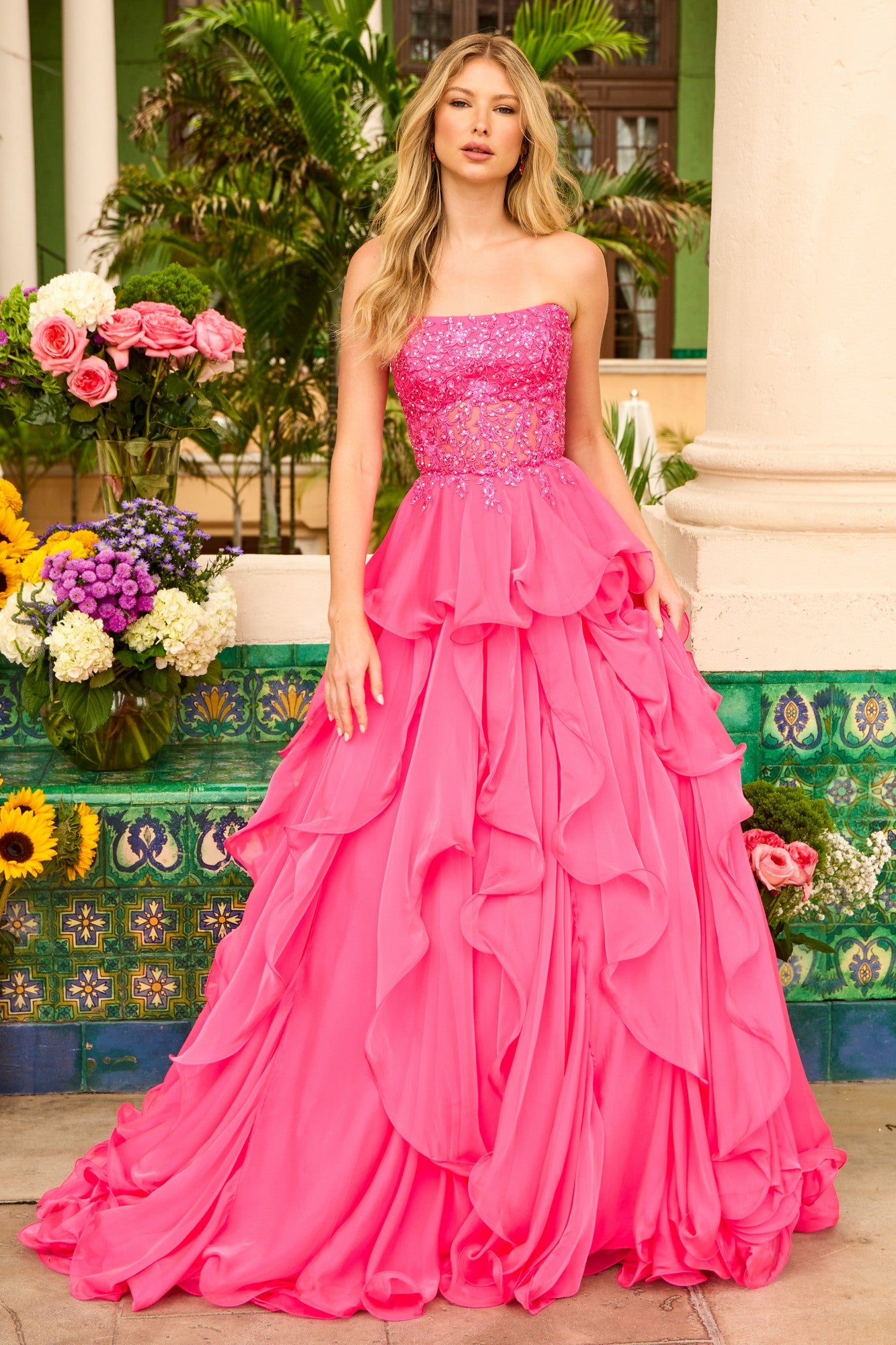 A woman wearing the Ava Presley 29133 Strapless Ruffle Ball Gown in bright pink stands outdoors by columns, surrounded by flowers and greenery.