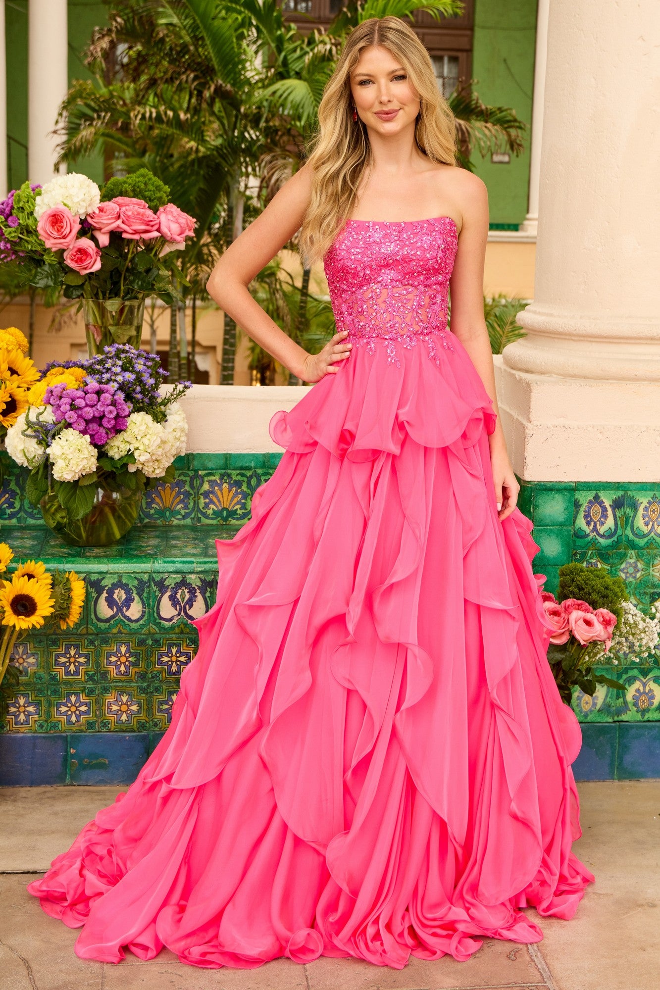 A woman stands outdoors in the strapless Ava Presley 29133 ball gown, featuring a bright pink ruffled skirt and an embroidered corset bodice, surrounded by colorful flowers and greenery.