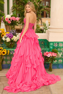 A woman stands outdoors amid flowers and greenery, wearing the Ava Presley 29133 Strapless Ruffle Ball Gown with a bright pink, layered skirt and a long, flowing train.