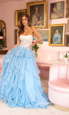 A woman stands in a pink room with floral arrangements and portrait paintings, wearing the Ava Presley 29133 Strapless Ruffle Ball Gown featuring an embroidered corset bodice and layered skirt.
