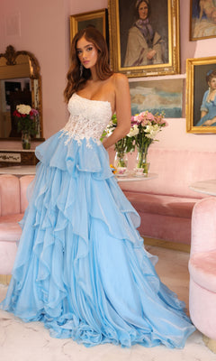 A woman wearing the Ava Presley 29133 Strapless Ruffle Ball Gown, featuring an embroidered corset bodice and flowing blue layered skirt, stands in a room with pink furniture, flowers, and framed portraits on the wall.