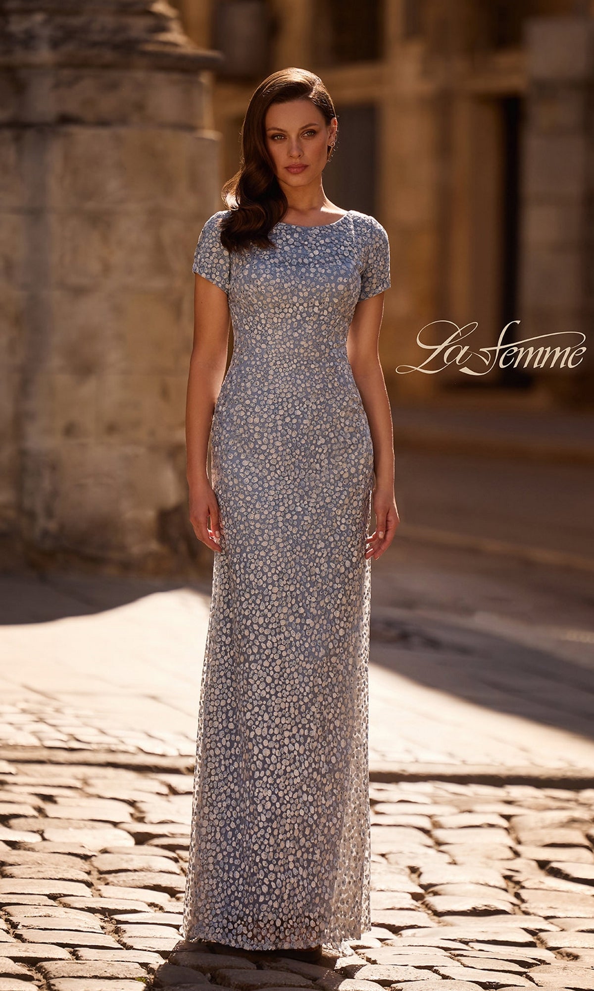 A woman in the Long Formal Dress: La Femme 29134, steel blue with rhinestone details and a fitted sheath silhouette, stands on a cobblestone street in soft sunlight.