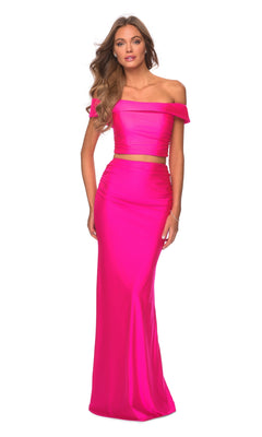A woman poses against a white background in the La Femme Bright Neon Two-Piece Prom Dress 2914, featuring an off-the-shoulder pink crop top and a matching high-waisted, floor-length skirt.