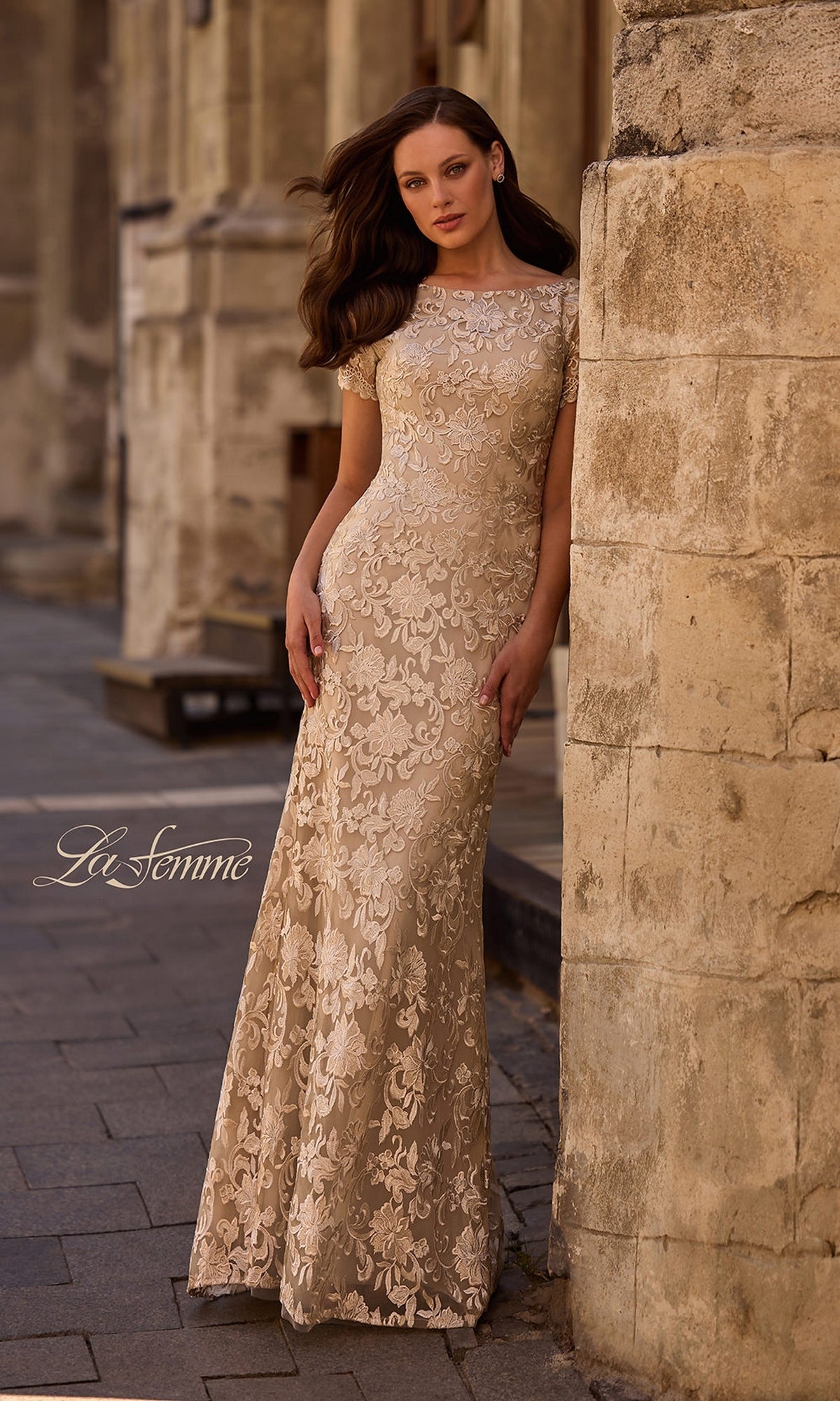 A woman in the La Femme 29161 long formal dress, a champagne-colored, floor-length sheath with short sleeves, stands against a stone building on a city sidewalk.