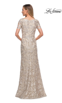 A woman is shown from the back wearing the La Femme 29161, a long formal, champagne-colored lace sheath dress with short sleeves and intricate floral embroidery. The “La Femme” logo appears in the top right corner. This elegant floor-length dress is an ideal choice for a mother-of-the-bride ensemble.