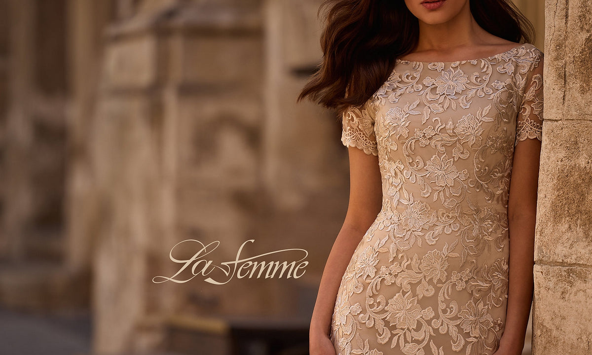 A woman wearing the La Femme 29161 long formal dress stands against a stone wall. This elegant beige lace sheath features floral embroidery and short sleeves, making it a perfect mother-of-the-bride choice. The words “La Femme” appear over the image.