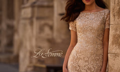 A woman wearing the La Femme 29161 long formal dress stands against a stone wall. This elegant beige lace sheath features floral embroidery and short sleeves, making it a perfect mother-of-the-bride choice. The words “La Femme” appear over the image.