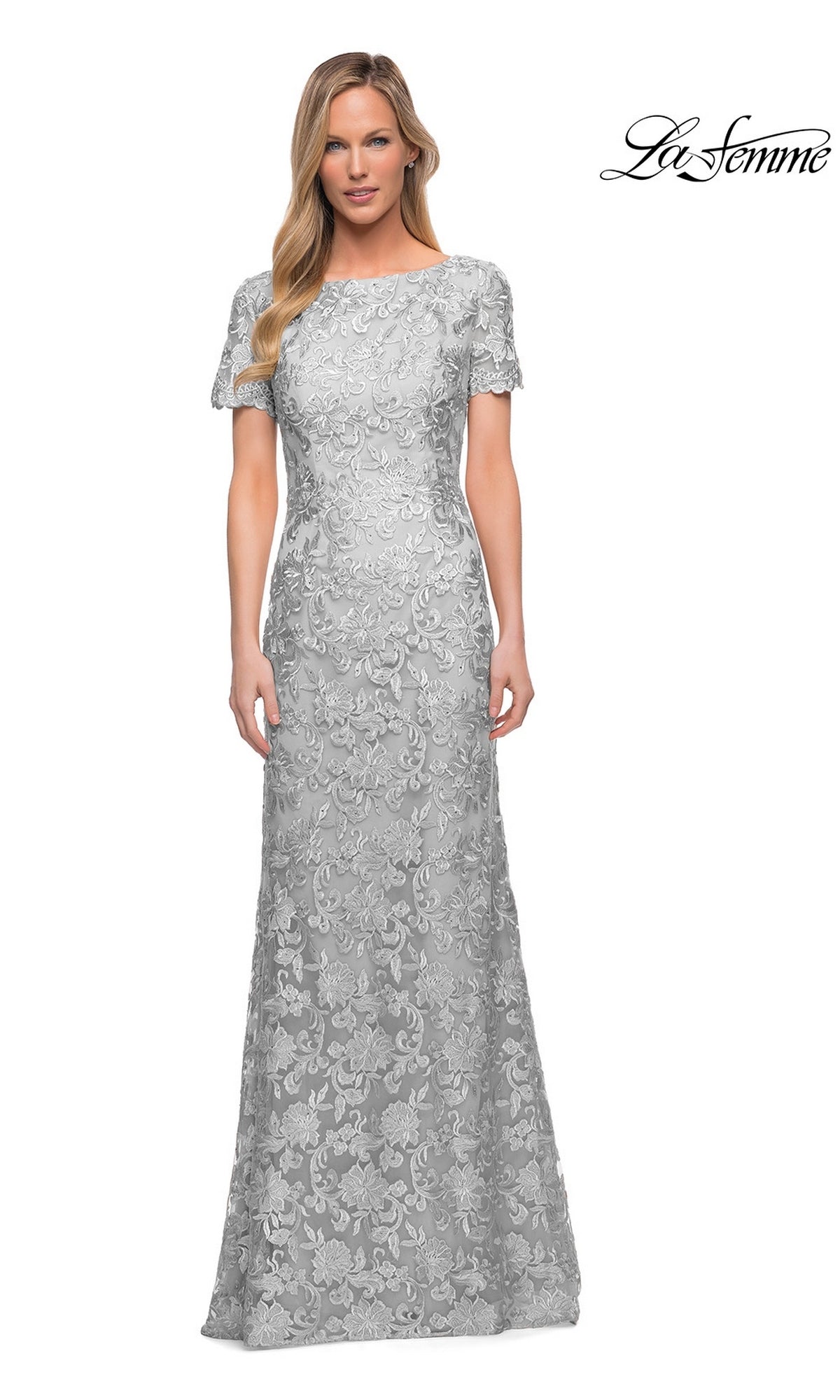 A woman is wearing the La Femme 29161 long formal dress—a floor-length, short-sleeve sheath gown in silver with floral lace patterns—standing against a plain white background.