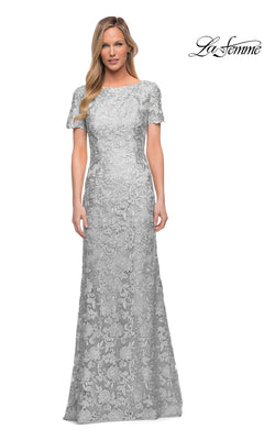 A woman is wearing the La Femme 29161 long formal dress—a floor-length, short-sleeve sheath gown in silver with floral lace patterns—standing against a plain white background.