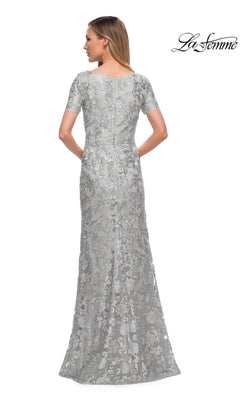 A woman is shown from the back against a white background with the “La Femme” logo in the top right corner, wearing a silver, floor-length, short-sleeved lace sheath known as the Long Formal Dress: La Femme 29161.