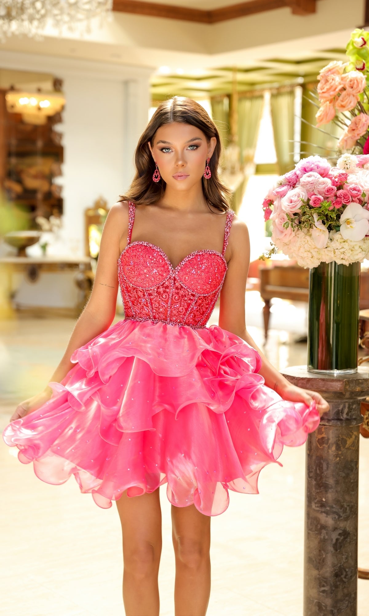 A young woman stands indoors in the Ava Presley 29179 Tiered Short Corset Hoco Dress, a bright pink ruffled dress with a beaded corset bodice, beside a large bouquet of flowers on a marble pedestal.