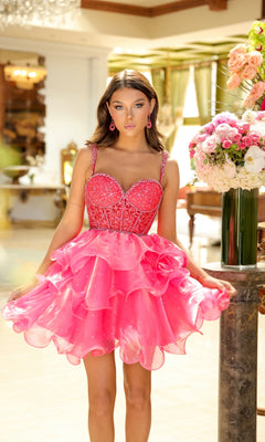 A young woman stands indoors in the Ava Presley 29179 Tiered Short Corset Hoco Dress, a bright pink ruffled dress with a beaded corset bodice, beside a large bouquet of flowers on a marble pedestal.