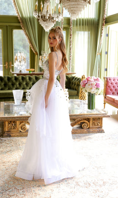 A woman in a white Tiered Long Prom Ball Gown, Ava Presley 29189, stands in an elegant room with green velvet sofas, chandeliers, and a large bouquet on a glass coffee table.
