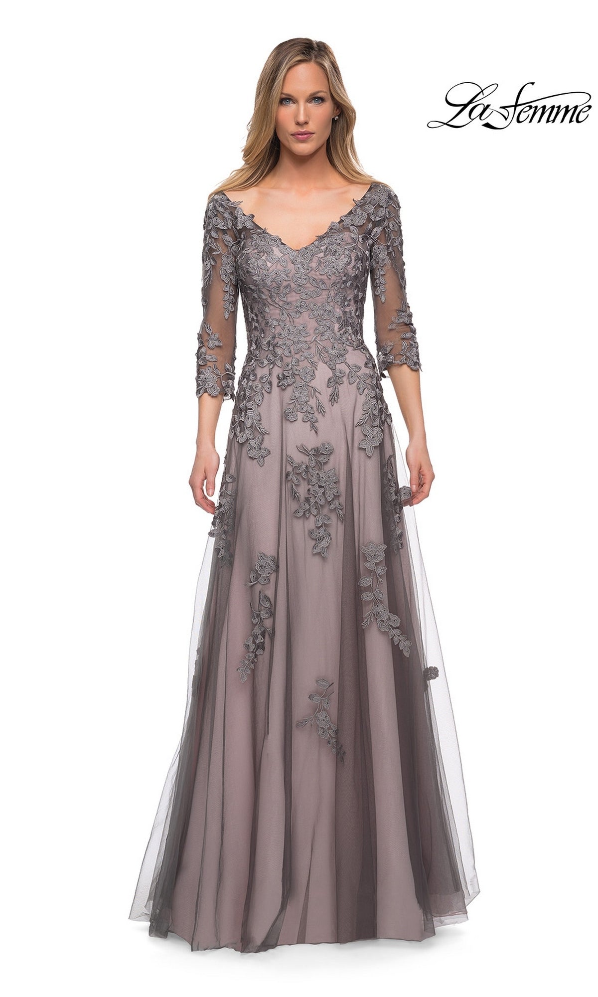 A woman wears the Long Formal Dress: La Femme 29205 in pink/gray, a floor-length A-line tulle gown featuring lace appliqué and three-quarter sleeves, while standing against a white background. The “La Femme” logo is displayed in the top right corner.