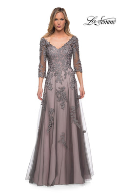 A woman wears the Long Formal Dress: La Femme 29205 in pink/gray, a floor-length A-line tulle gown featuring lace appliqué and three-quarter sleeves, while standing against a white background. The “La Femme” logo is displayed in the top right corner.