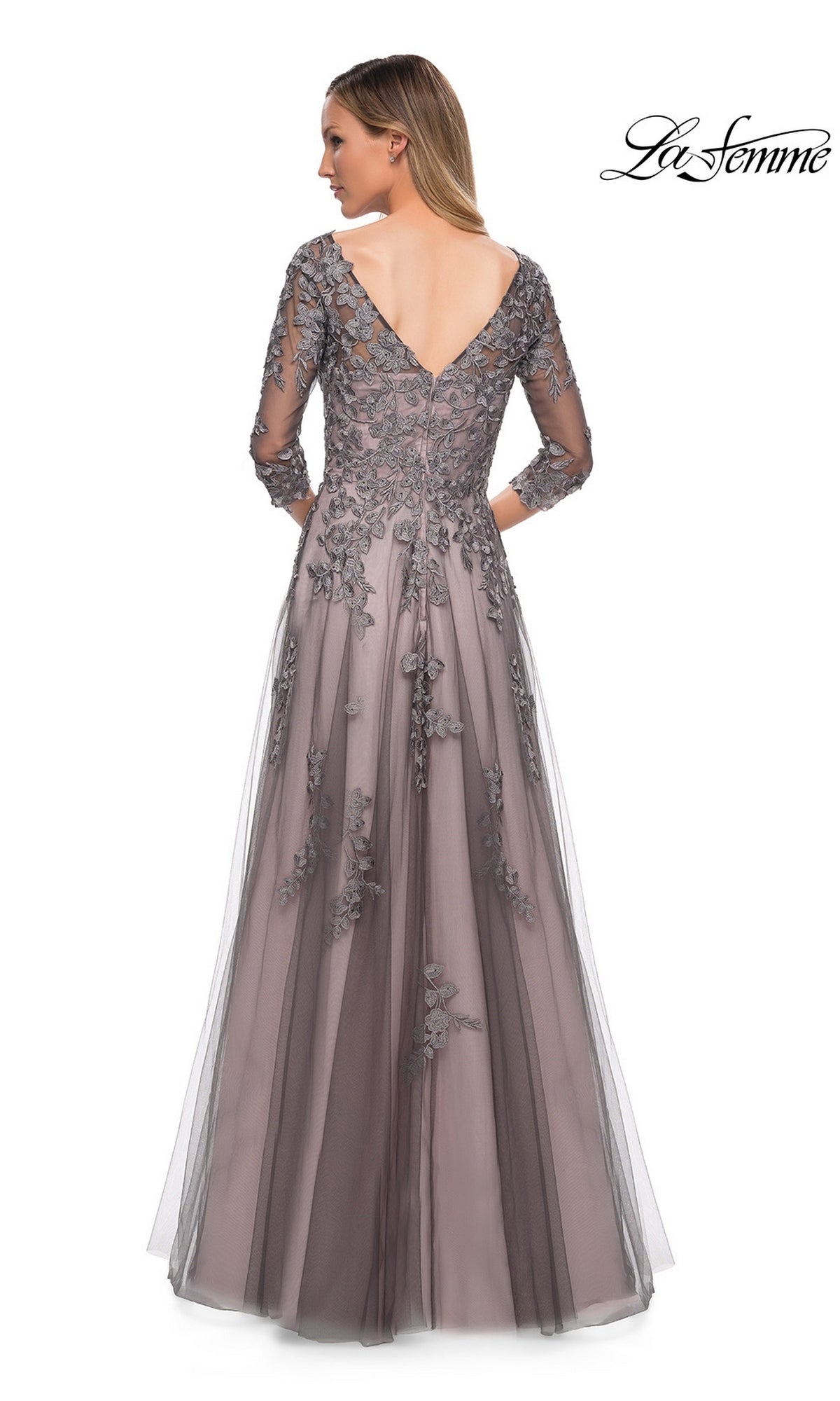 A woman wearing the La Femme 29205 long formal dress stands with her back to the camera, showcasing its delicate floral lace accents and V-shaped back. The elegant A-line tulle gown flows gracefully in soft grey, while the “La Femme” logo appears in the top right corner.