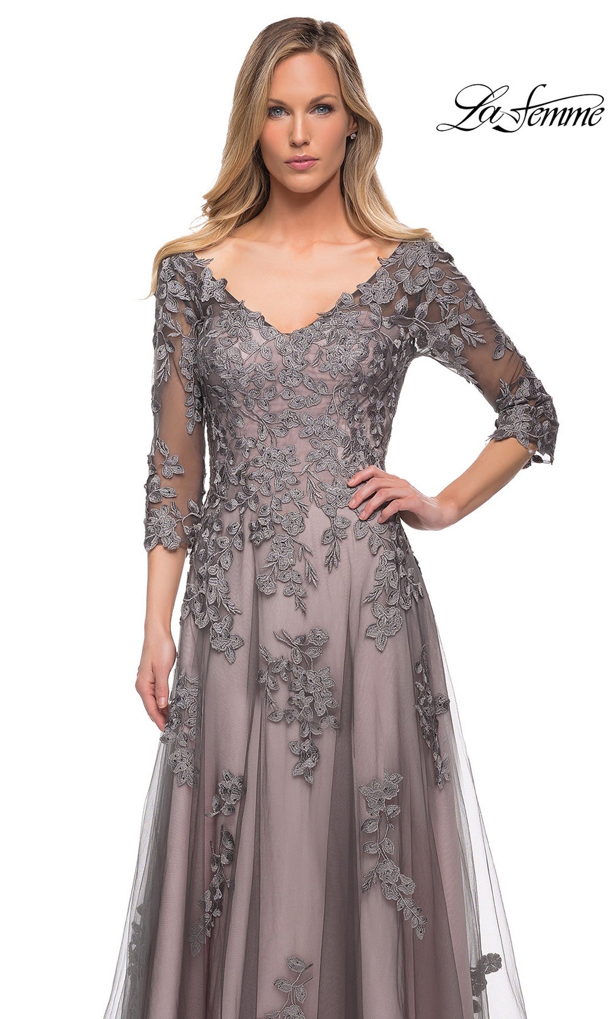 A woman wearing the Long Formal Dress: La Femme 29205—a taupe, long A-line tulle gown featuring gray floral lace appliqué, three-quarter sleeves, and a scalloped neckline. The “La Femme” logo appears in the top right corner. The background is plain white.