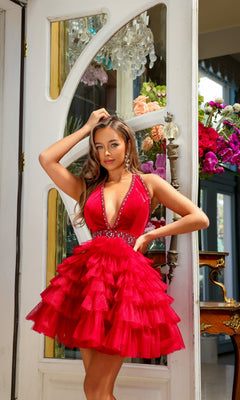 A woman stands indoors by a door, posing with one hand on her hip and the other touching her hair, wearing the V-Neck Short Halter Ruffled Homecoming Dress 29206 in red.