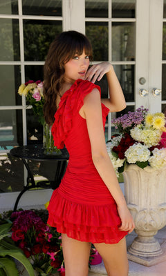 A woman stands outdoors near a glass door, surrounded by colorful flowers, wearing the Ruffled One-Shoulder Short Homecoming Dress 29212 in red.