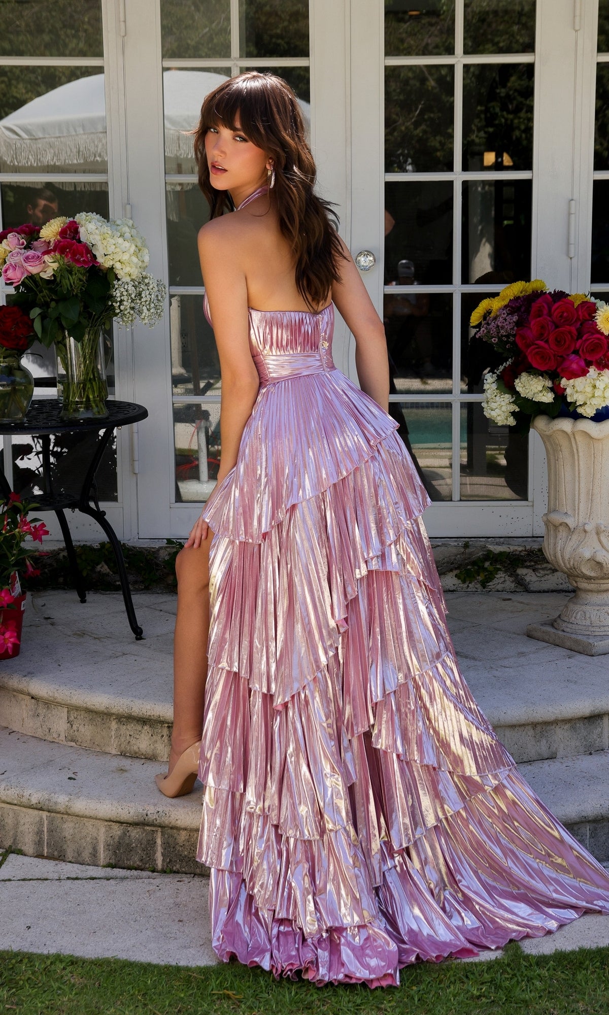 A woman in the Long Prom Dress 29220 by Ava Presley, featuring a metallic bodice and tiered pink skirt, poses outdoors on stone steps with her back slightly turned, surrounded by large floral arrangements.