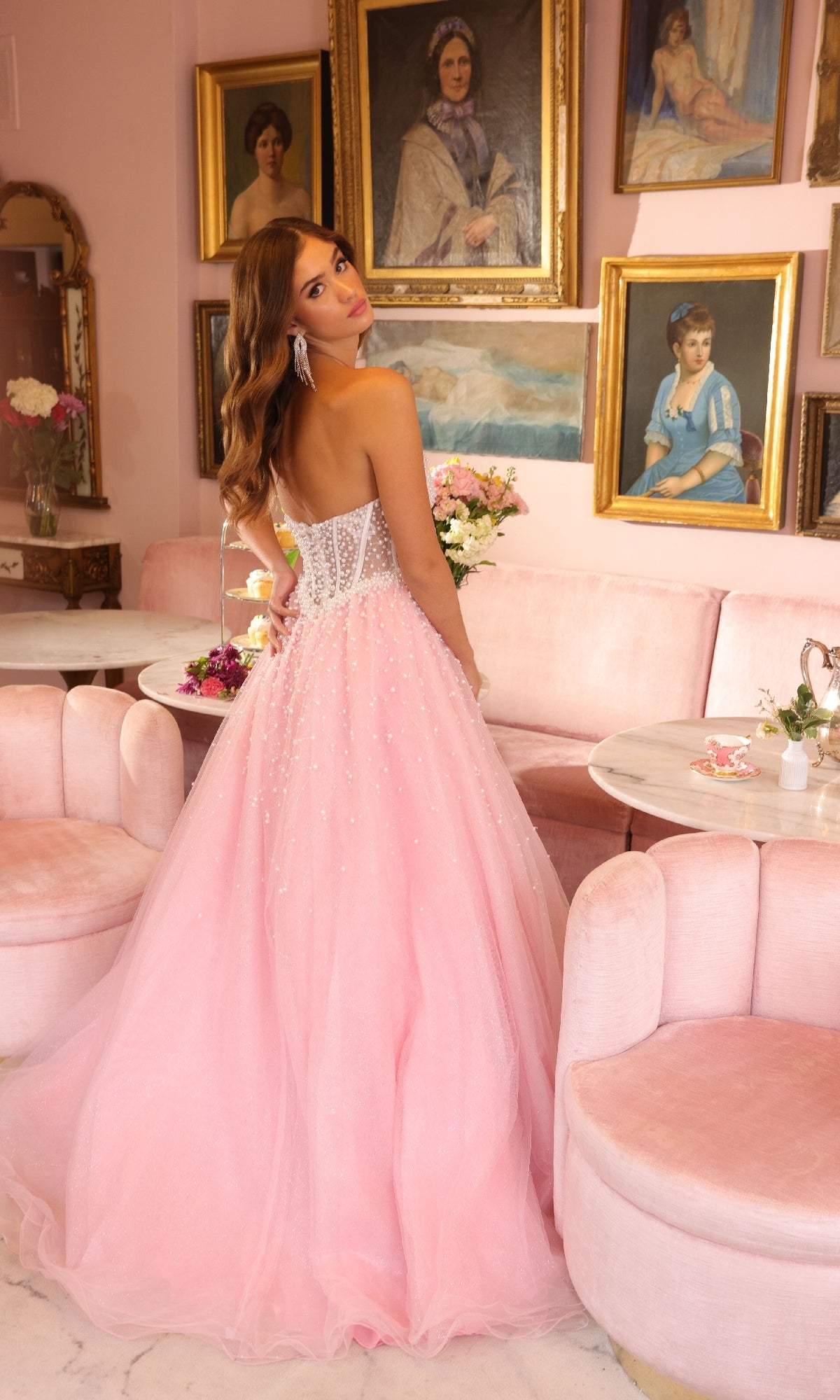 A woman in the Ava Presley 29226 Corset Tulle Strapless Ball Gown with pearl embellishments stands in a pink-themed room with vintage paintings and velvet furniture, looking over her shoulder at the camera.