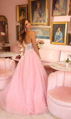 A woman in the Ava Presley 29226 Corset Tulle Strapless Ball Gown with pearl embellishments stands in a pink-themed room with vintage paintings and velvet furniture, looking over her shoulder at the camera.