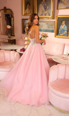 A woman wears the Ava Presley 29226 corset tulle strapless ball gown with pearl accents and a lace-up back, standing in an elegant room with pink furniture and framed portraits on the walls.