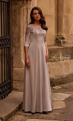A woman in the Long Formal Dress: La Femme 29227 in silver, featuring lace detailing and an A-line silhouette, stands outside near a stone building.