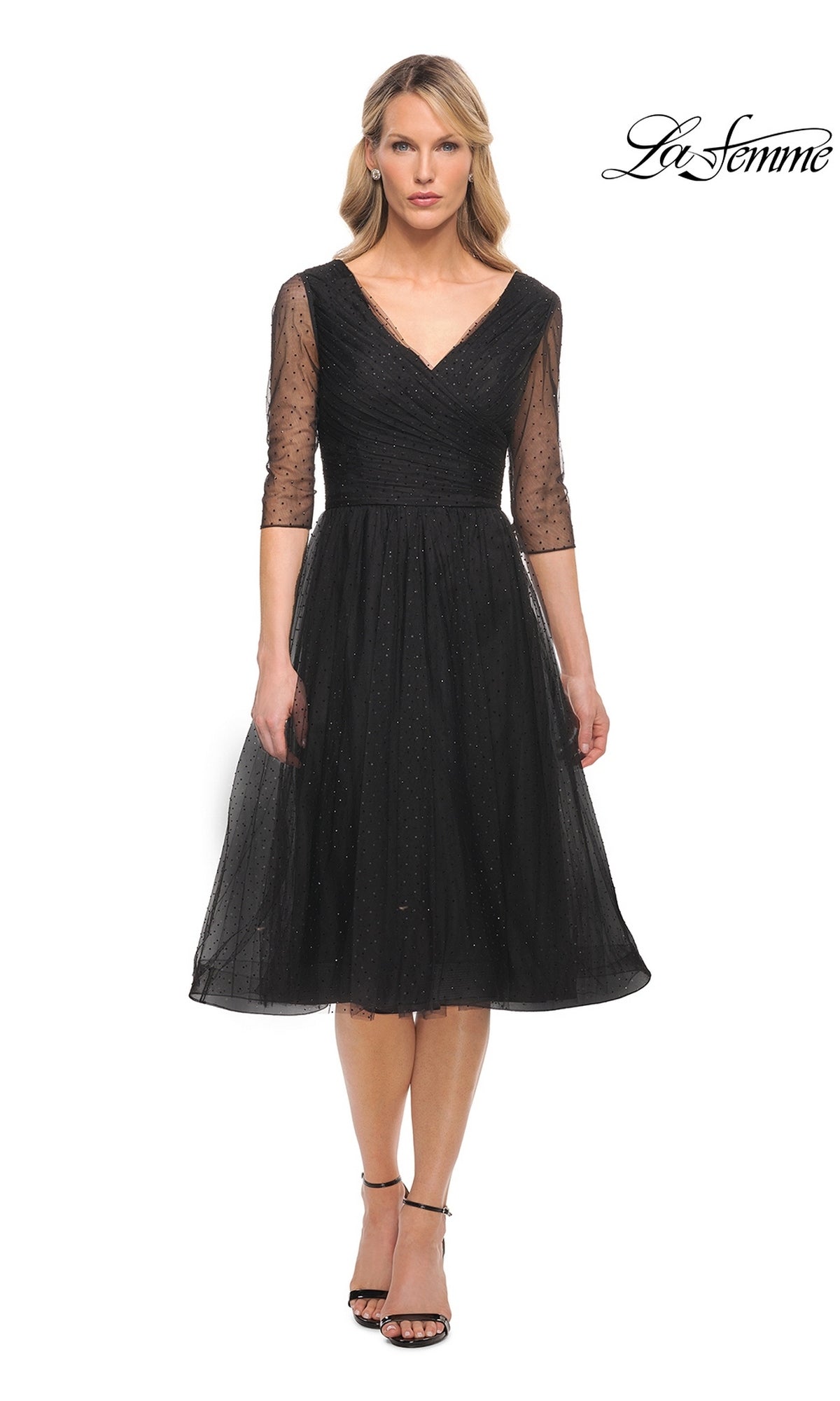 Woman in a black Knee Length Wedding Guest Dress: La Femme 29229 with sheer three-quarter sleeves and V-neckline, standing against a white background. “La Femme” logo top right.