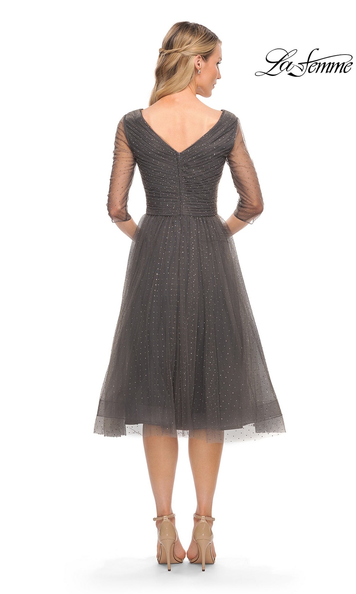 Woman in Knee Length Wedding Guest Dress: La Femme 29229, featuring sheer sleeves, sparkling embellishments, V-back, and fit-and-flare silhouette, stands with her back to the camera.
