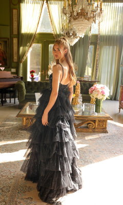 A woman in the Long Prom Dress 29232 by Ava Presley—a formal black a-line gown with ruffles and a sweetheart neckline—stands in an elegant, sunlit room with large windows, chandeliers, and ornate furnishings.