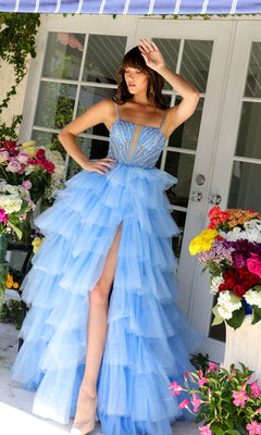 Wearing the Long Prom Dress 29232 by Ava Presley, a woman stands on a porch surrounded by colorful flowers, raising one hand to shield her eyes. The gown features light blue layers, ruffles, an A-line silhouette, and a high slit.