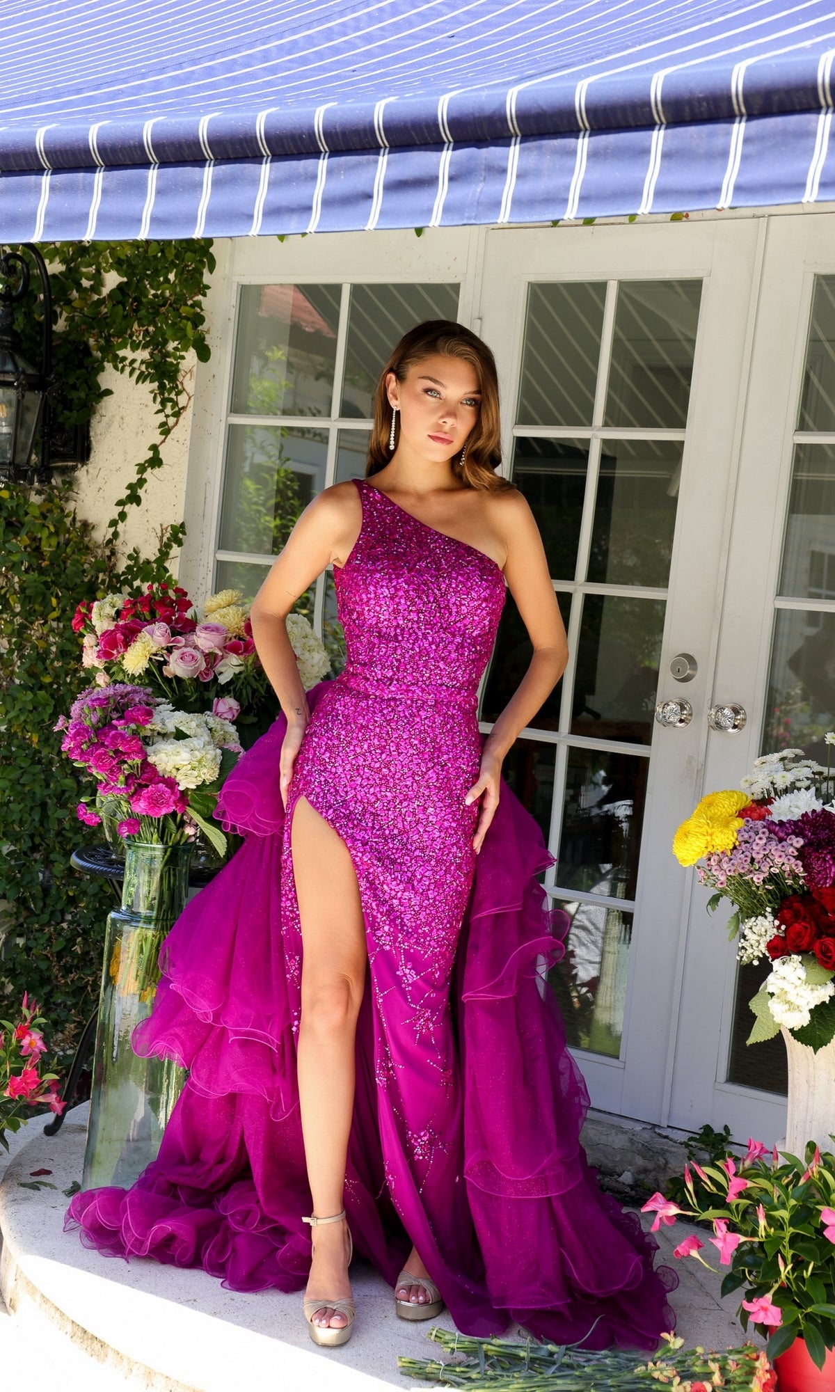 A woman stands on a porch, surrounded by colorful flower arrangements, wearing the Ava Presley 29236 one-shoulder beaded prom dress with a high slit in bright magenta.