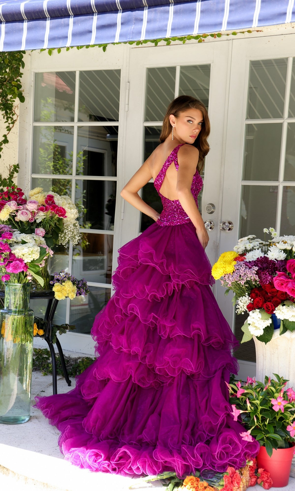 A woman in a ruffled, bright purple One-Shoulder Beaded Prom Dress: Ava Presley 29236 stands outside among vibrant flowers, facing away from white-framed glass doors.