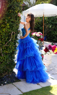 A woman wearing the One-Shoulder Beaded Prom Dress: Ava Presley 29236 stands outdoors beside a green hedge, with colorful flower arrangements and a white umbrella in the background.