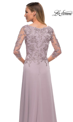 A woman wears the Long Formal Dress: La Femme 29251, a light purple lace dress with sheer, floral embroidered sleeves and back, facing away. The “La Femme” logo is top right.