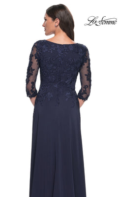 A woman in a navy lace Long Formal Dress: La Femme 29251 with sheer sleeves and floral embroidery stands before a white background. The logo appears in the top right corner.