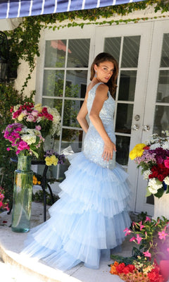 A woman in the Ava Presley 29254 long formal dress, a light blue ruffled one-shoulder evening gown, stands before glass double doors, surrounded by colorful flower arrangements.