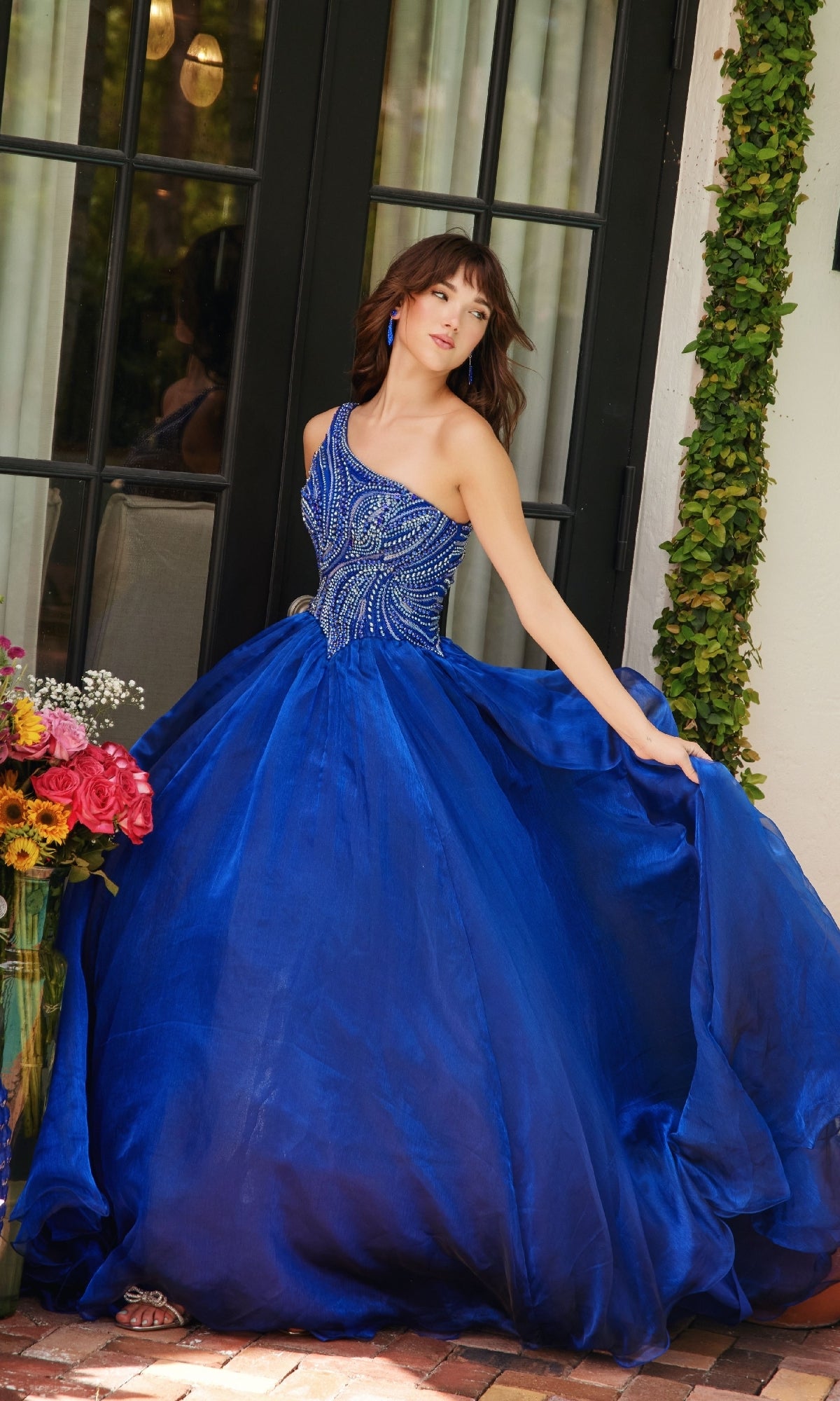 A woman stands near a glass door holding her Ava Presley 29258 one-shoulder prom ball gown with a beaded bodice; a colorful bouquet of flowers rests nearby.