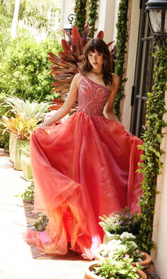 Wearing the Ava Presley 29258 One-Shoulder Prom Ball Gown, a woman stands on a brick walkway among lush plants and flowers, holding out the skirt of the vibrant pink and orange beaded dress.