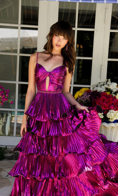 A person poses outdoors in front of glass doors, wearing the Ava Presley 29263 Long Metallic Ruffle Prom Dress, featuring a shiny magenta color and tiered skirt, with a beaded waist and a bouquet of flowers nearby.