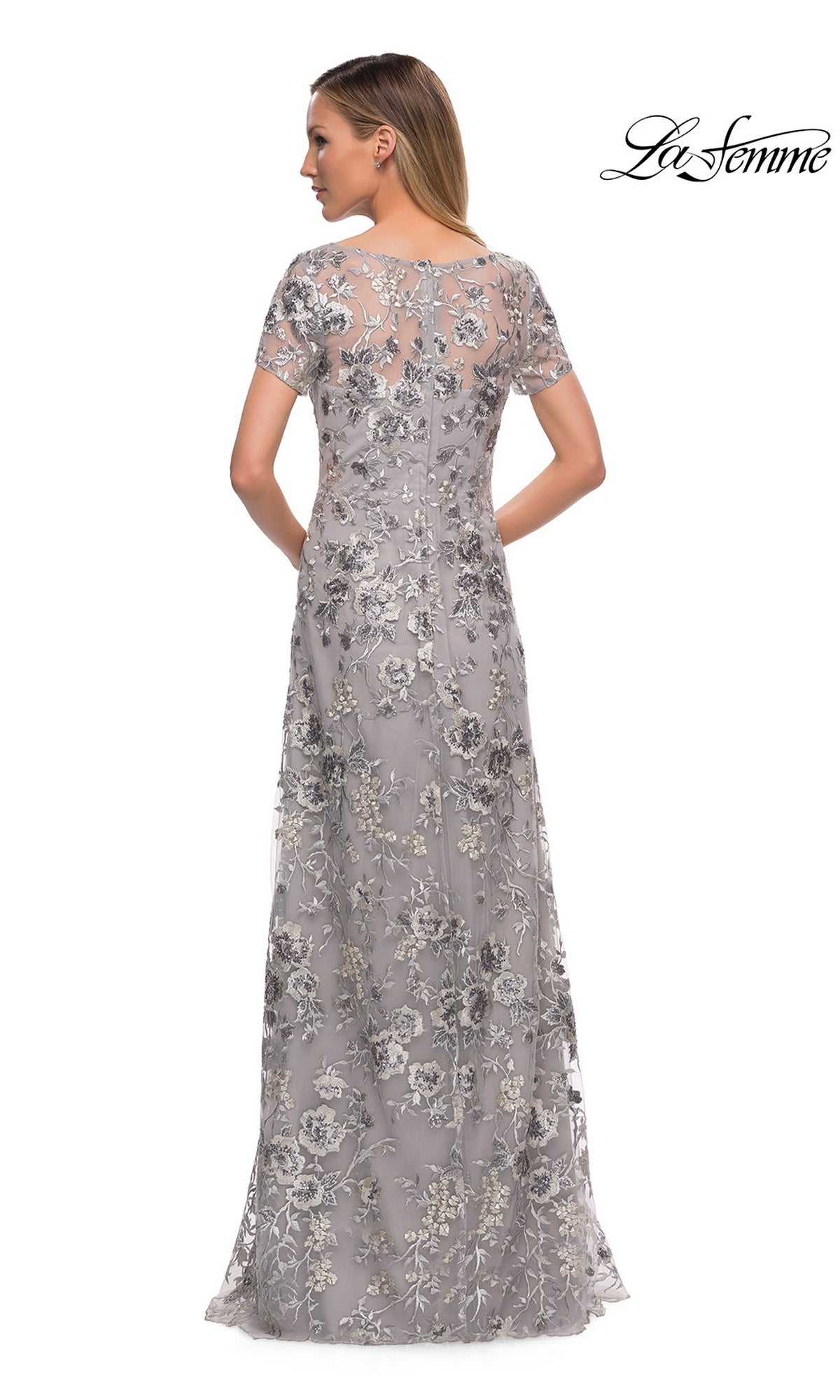 A woman wears the Long Formal Dress: La Femme 29281, a floor-length grey modest dress with short sleeves and silver floral embroidery.