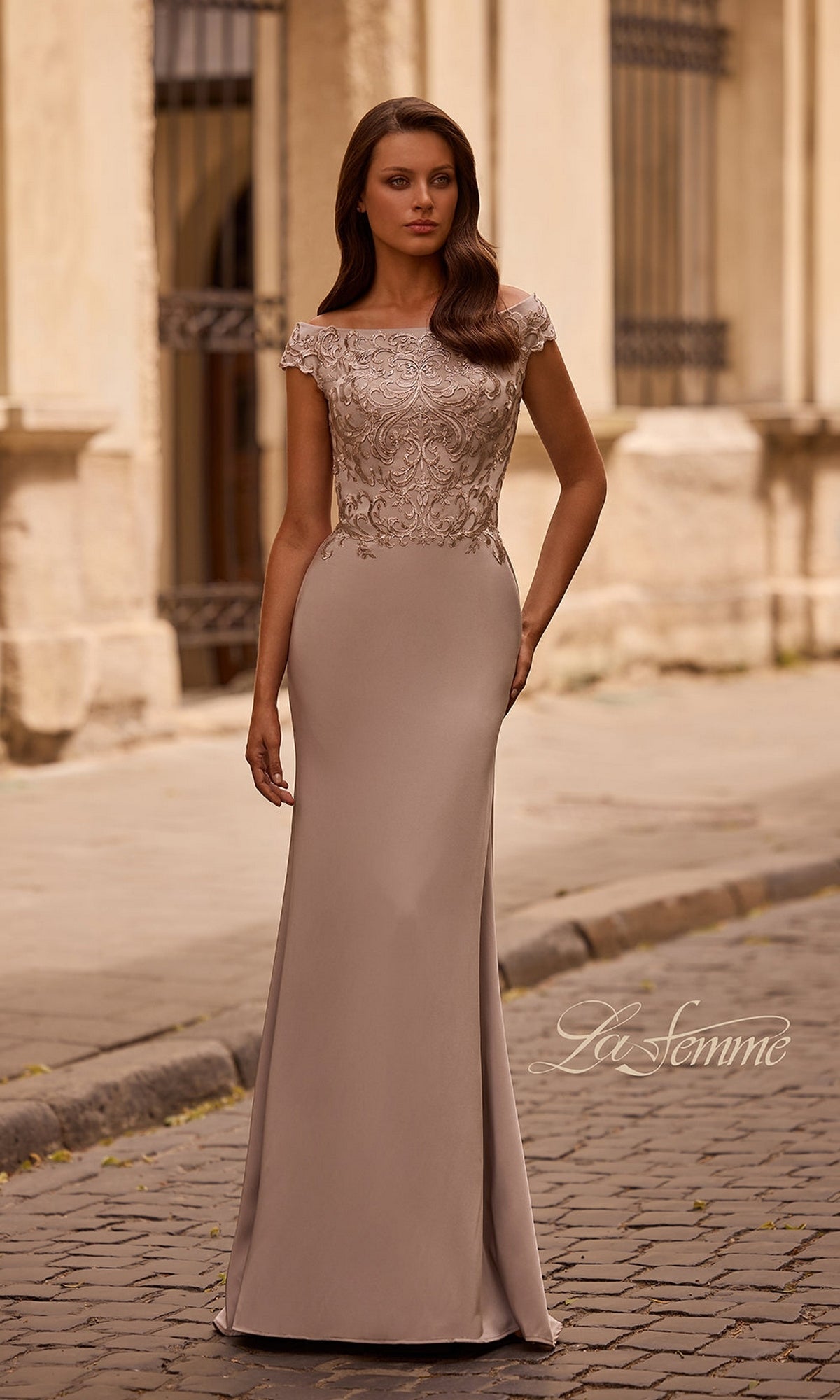 Woman wearing Long Formal Dress: La Femme 29331 in light taupe, with a fitted embroidered bodice and cap sleeves, stands on a cobblestone street by a light-colored building.