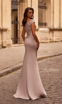 A woman in the Long Formal Dress: La Femme 29331, featuring a fitted embroidered bodice and off-the-shoulder neckline, stands on a cobblestone street looking back.