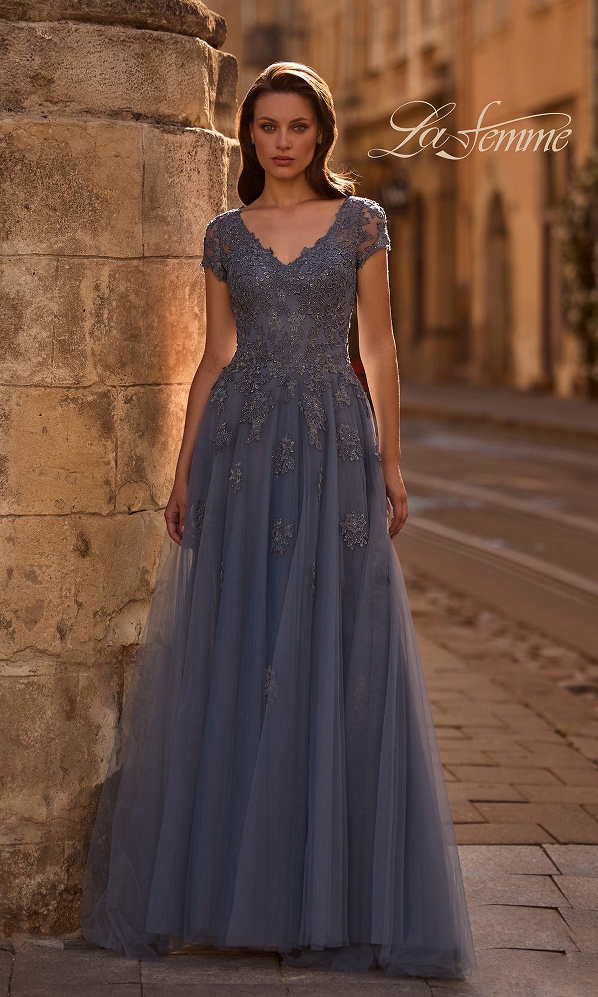 A woman in the Long Formal Dress: La Femme 29380 in slate poses by a stone column on a cobblestone street, with “La Femme” text in the background.