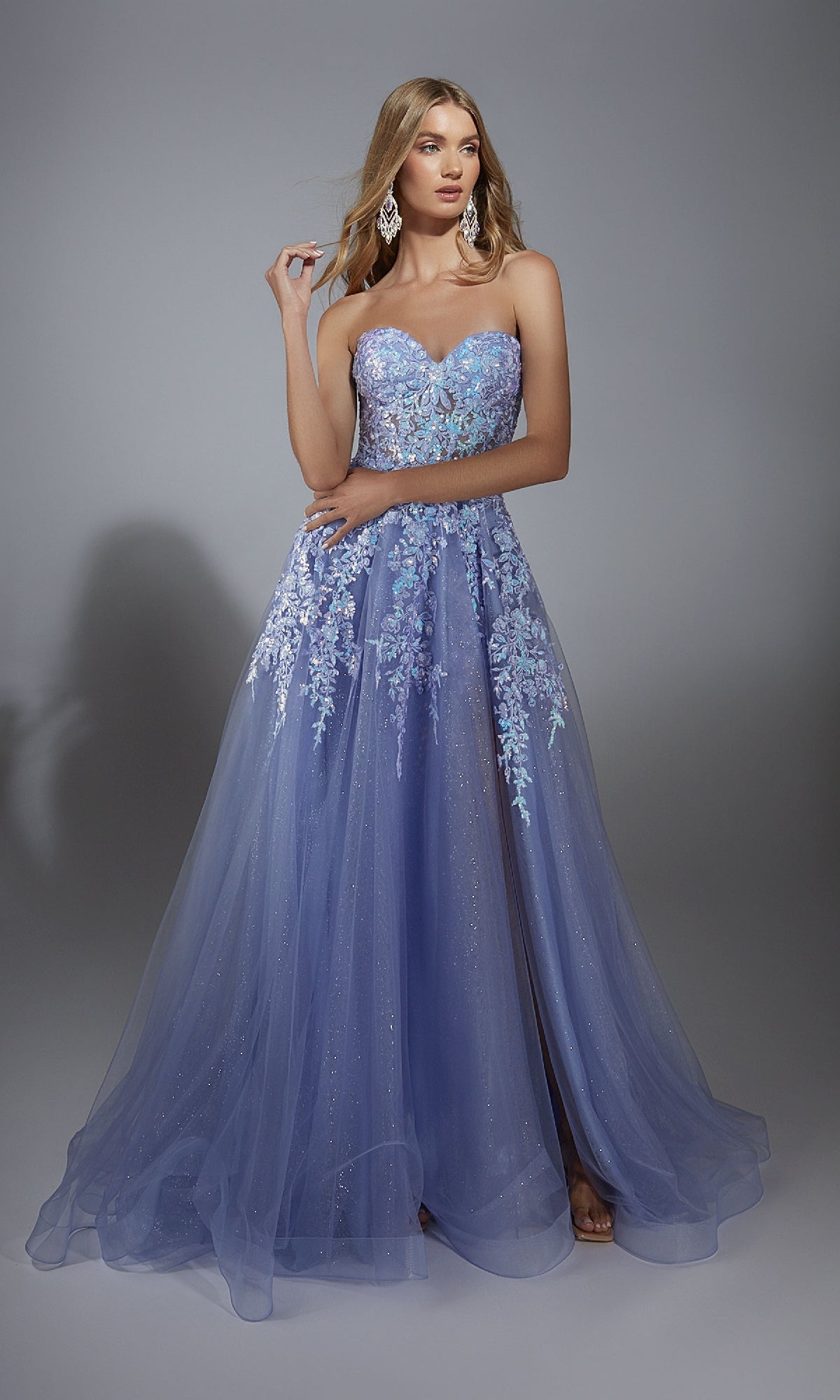 A woman stands against a plain gray backdrop wearing the Aerie Couture 293: a strapless glitter prom ball gown with floral lace embroidery and a flowing tulle skirt in lavender.