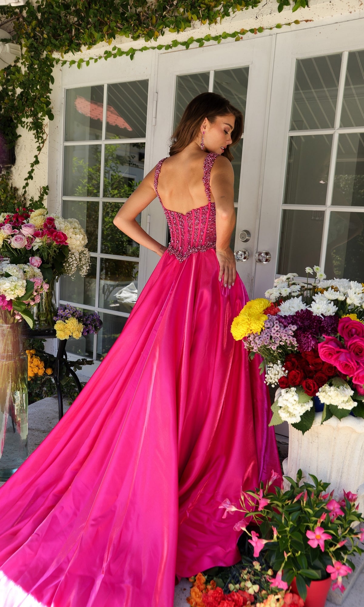 A woman stands with her back to the camera, surrounded by vibrant flowers outside glass doors, wearing the Long Formal Dress 29521 by Ava Presley—a bright pink, floor-length gown with a beaded corset bodice.