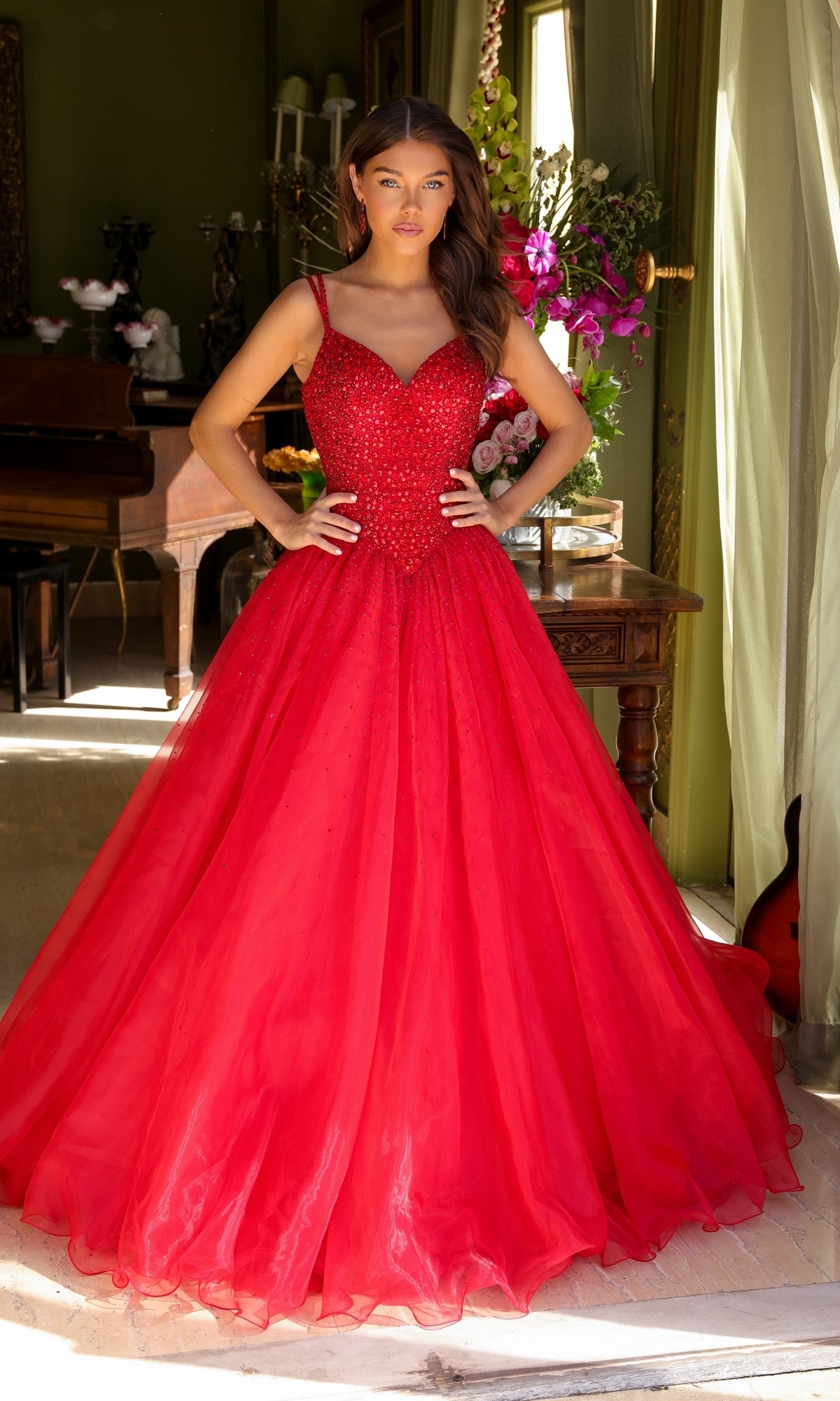 A woman stands indoors wearing the Long Prom Dress 29522 by Ava Presley, a floor-length, bright red A-line gown with a fitted, beaded bodice and thin straps. She poses with hands on hips amidst floral decor.