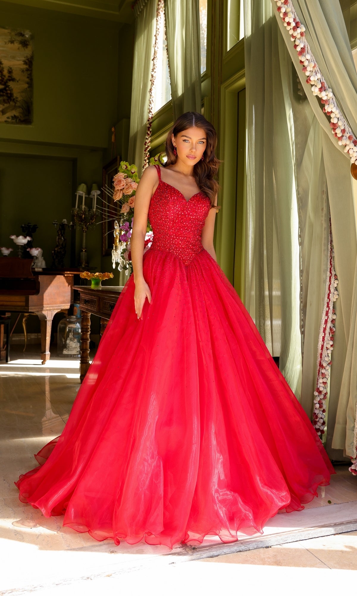 A woman wears the Long Prom Dress 29522 by Ava Presley, an A-line formal gown with a sweetheart neckline and beaded bodice, standing indoors near large windows with green curtains and floral decor.