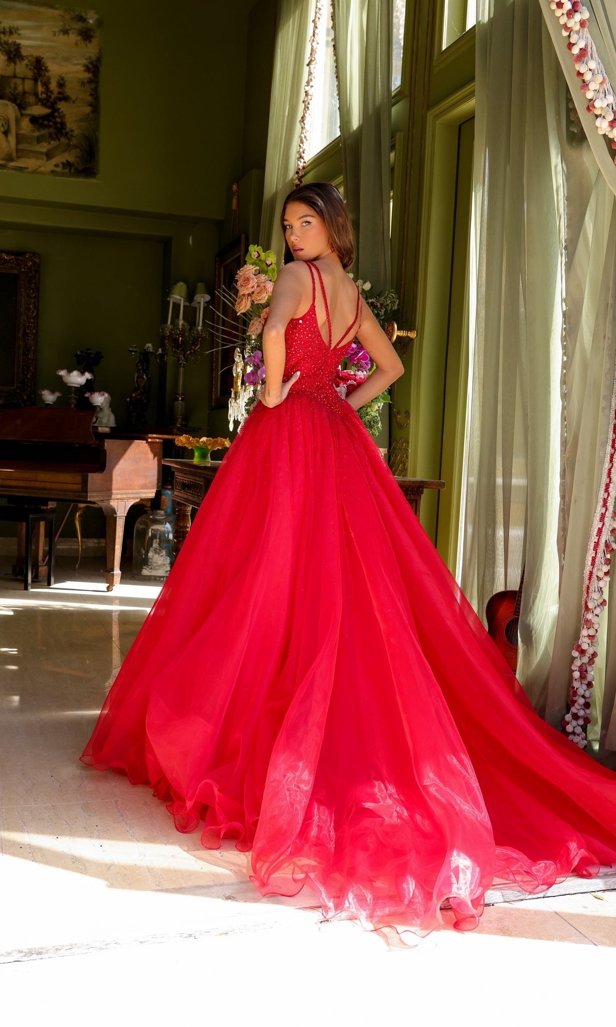 A woman poses indoors by a large window and elegant floral arrangements, wearing the Long Prom Dress 29522 by Ava Presley—a bright red A-line formal gown with train, looking over her shoulder.